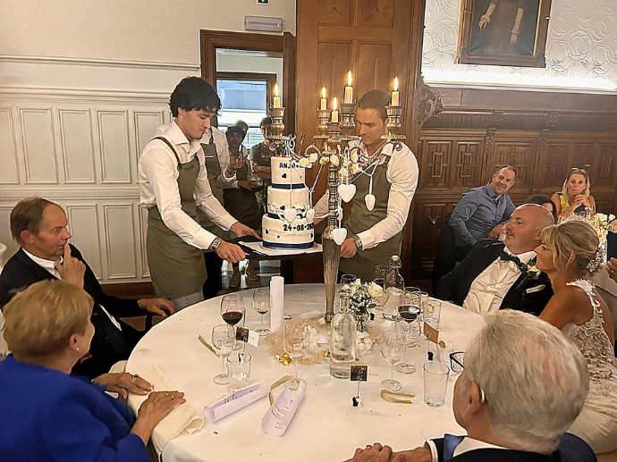 Bruidspaar en obers bij het aansnijden van een witte bruidstaart aan een ronde tafel.
